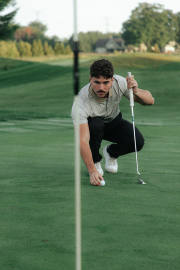 A man crouches on a golf green holding a putter and a golf ball, wearing a light beige country cactus patterned t-shirt, black pants, and white sneakers.