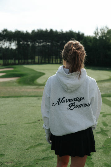 A woman standing on a golf course, viewed from behind, wearing a light grey hoodie with the words 'Normalize Bogeys' printed in black script across the back, paired with a black skirt.