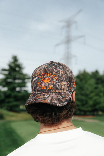 A man viewed from behind wearing a camouflage baseball cap with orange embroidered text that reads 'FLUSHED GOLF' and a white t-shirt, set against a blurred outdoor background of trees and a power line.