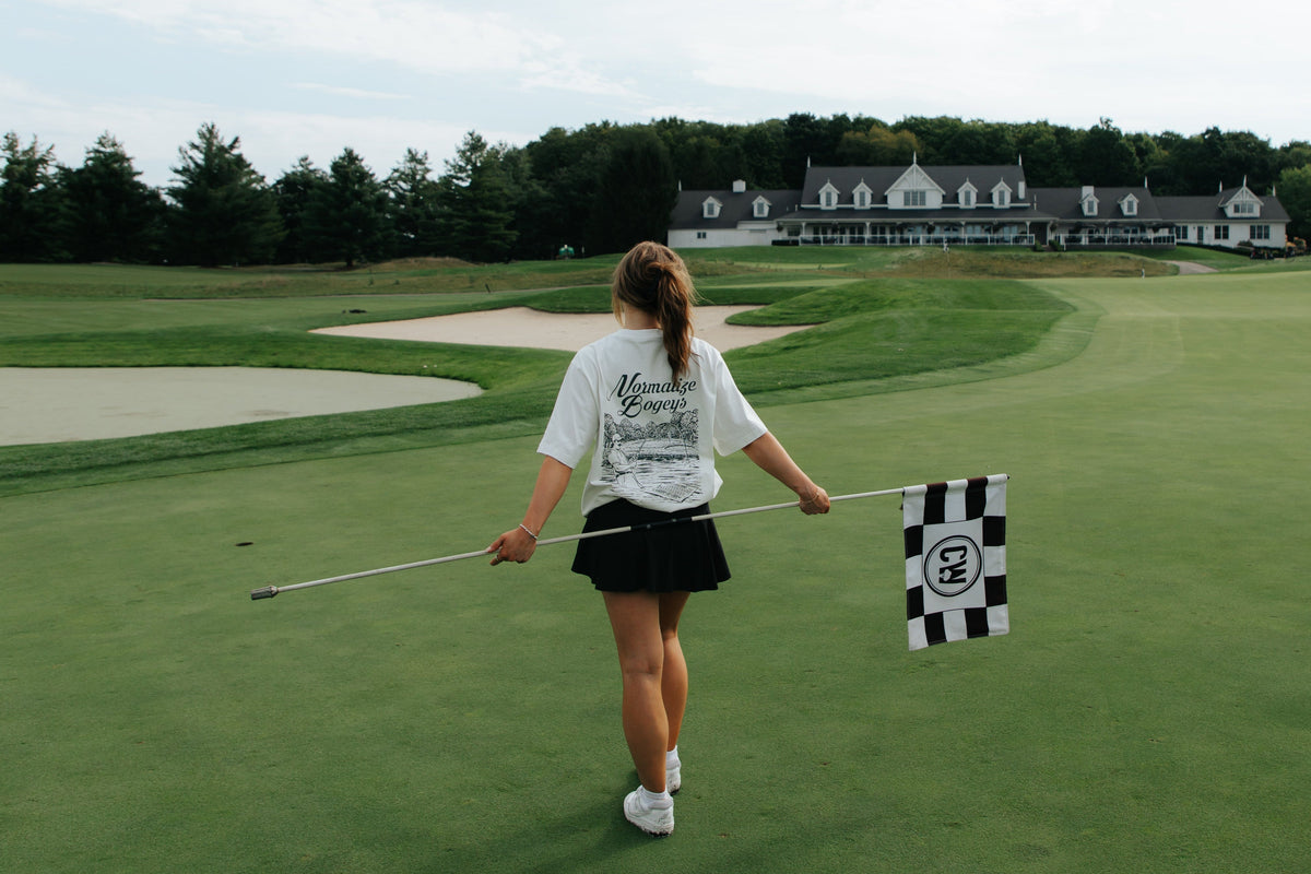 A woman on a golf course holds a checkered flagstick while walking toward a clubhouse in the distance under a cloudy sky.