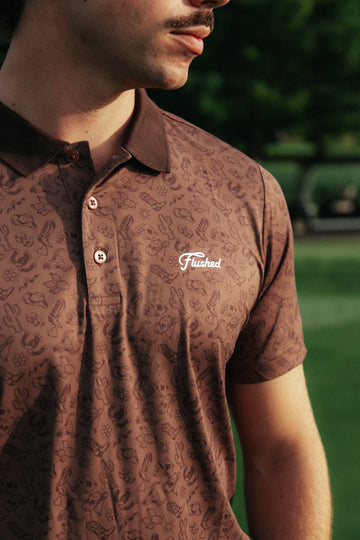 A close-up of a man wearing a brown short-sleeve polo shirt with an all-over country pattern and a white 'Flushed' logo on the chest, set against a blurred green outdoor background.
