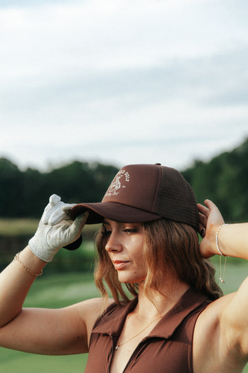 A woman on a golf course wearing a brown trucker-style cap with a white embroidered logo, a brown sleeveless top, and a white golf glove, adjusting her cap.