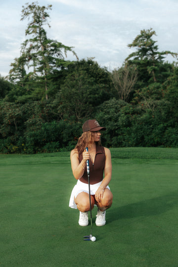 A woman crouches on a golf green holding a putter, wearing a brown golf bodysuit, white skirt, brown hat, and white sneakers, with trees in the background.
