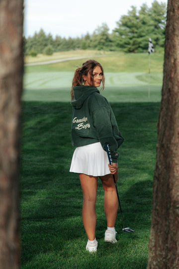 A woman stands on a golf course wearing a dark green cropped hoodie with 'normalize bogeys' written in white script on the back, a white pleated mini skirt, and white golf shoes, holding a golf club.