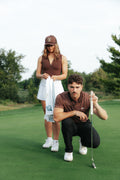 A man and a woman on a golf course using a towel to clean their golf balls . The man in the foreground is crouching and holding a golf club, wearing a brown patterned polo shirt, black pants, and white shoes.