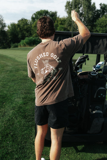 A man on a golf course seen from behind, wearing a brown t-shirt with a white graphic and the words 'FLUSHED GOLF country club' on the back, standing next to a golf cart.