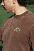A close-up side profile of a man wearing a brown washed t-shirt with a white circular graphic on the left chest that reads 'flushed golf country club'.
