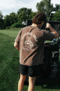 A man on a golf course stands with his back to the camera, wearing a brown t-shirt with a white graphic that reads 'FLUSHED GOLF Country Club' and black shorts, holding a golf club next to a cart.