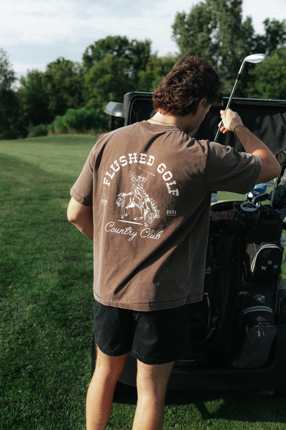 A man on a golf course stands with his back to the camera, wearing a brown t-shirt with a white graphic that reads 'FLUSHED GOLF Country Club' and black shorts, holding a golf club next to a cart.