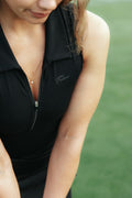 Close-up of a woman wearing a black sleeveless golf bodysuit polo zip-front top with a flushed golf script logo on the chest, photographed outdoors against a blurred green background.