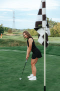 A woman in a black sleeveless bodysuit  polo and black pleated skirt plays golf on a green, holding a putter near a checkered flag in the foreground.