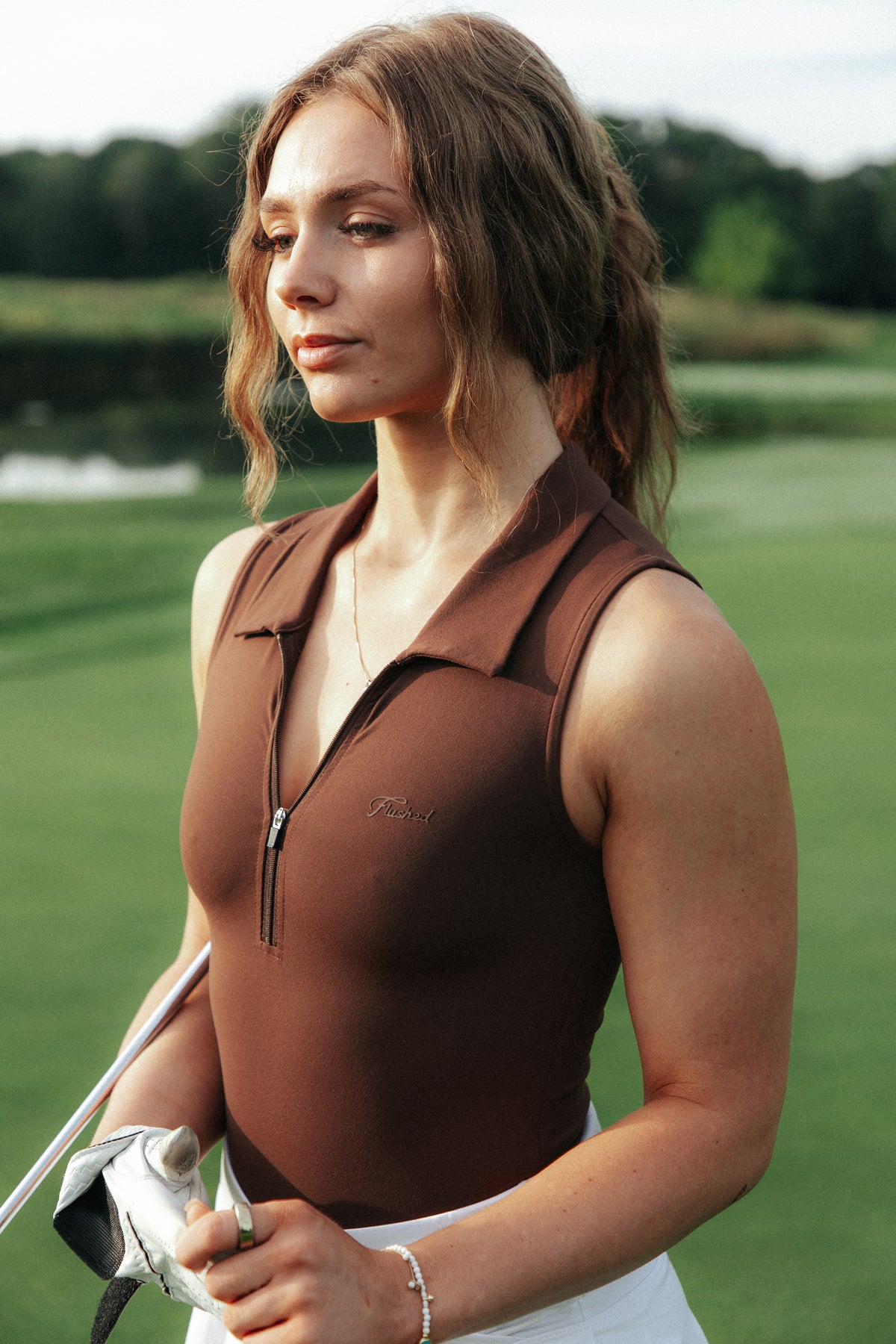A woman on a golf course wearing a fitted sleeveless brown golf bodysuit polo with a collar and zipper, paired with a white golf skirt, holding a golf club.
