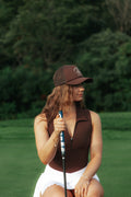 A woman on a golf course wearing a brown sleeveless bodysuit polo, a white skirt, and a brown flushed golf trucker cap, holding a golf club.