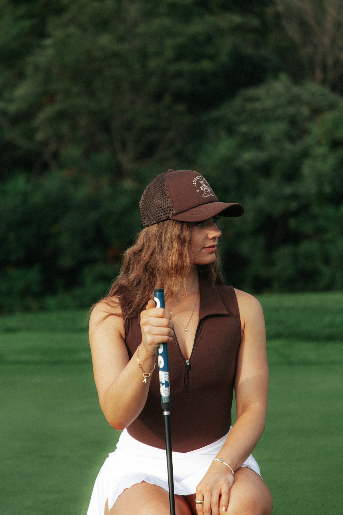 A woman on a golf course wearing a brown sleeveless bodysuit polo, a white skirt, and a brown flushed golf trucker cap, holding a golf club.