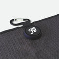 A close-up of a black circular carabiner clip on a golf waffle towel with a white stylized 'FG' logo, attached to a black strap and placed on a dark textured fabric surface.