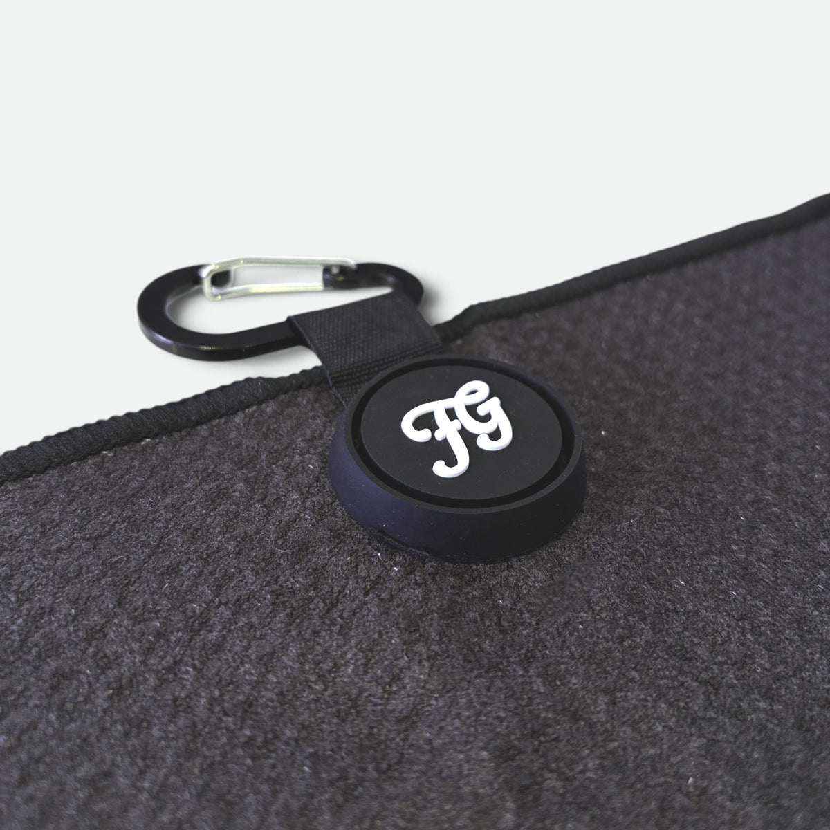 A close-up of a black circular carabiner clip on a golf waffle towel with a white stylized 'FG' logo, attached to a black strap and placed on a dark textured fabric surface.