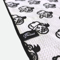 A close-up of a waffle golf towel with a repeating flushed golf mascot pattern and a black trim edge. A small black fabric tag with the word 'Flushed' in white script is sewn onto the edge.