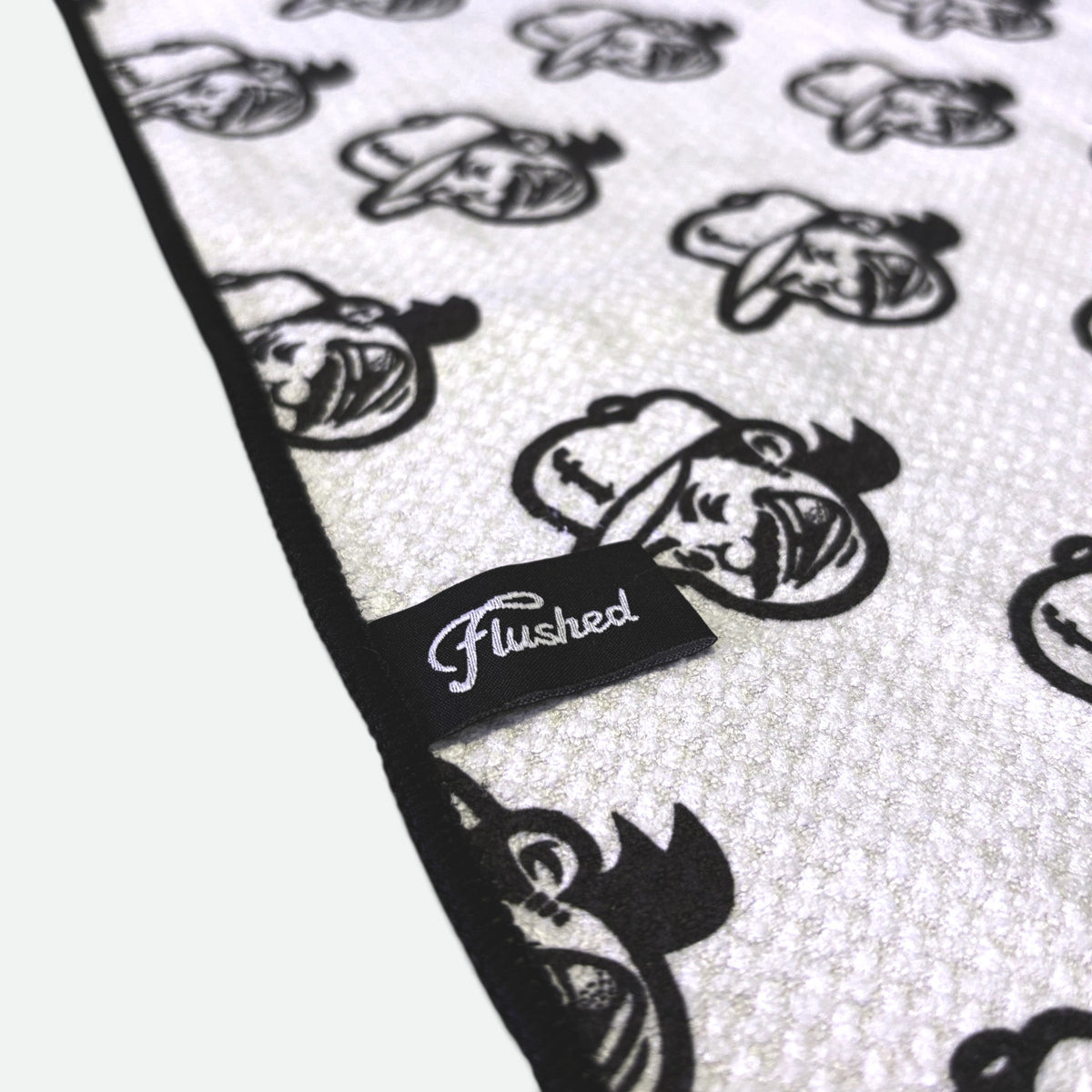 A close-up of a waffle golf towel with a repeating flushed golf mascot pattern and a black trim edge. A small black fabric tag with the word 'Flushed' in white script is sewn onto the edge.