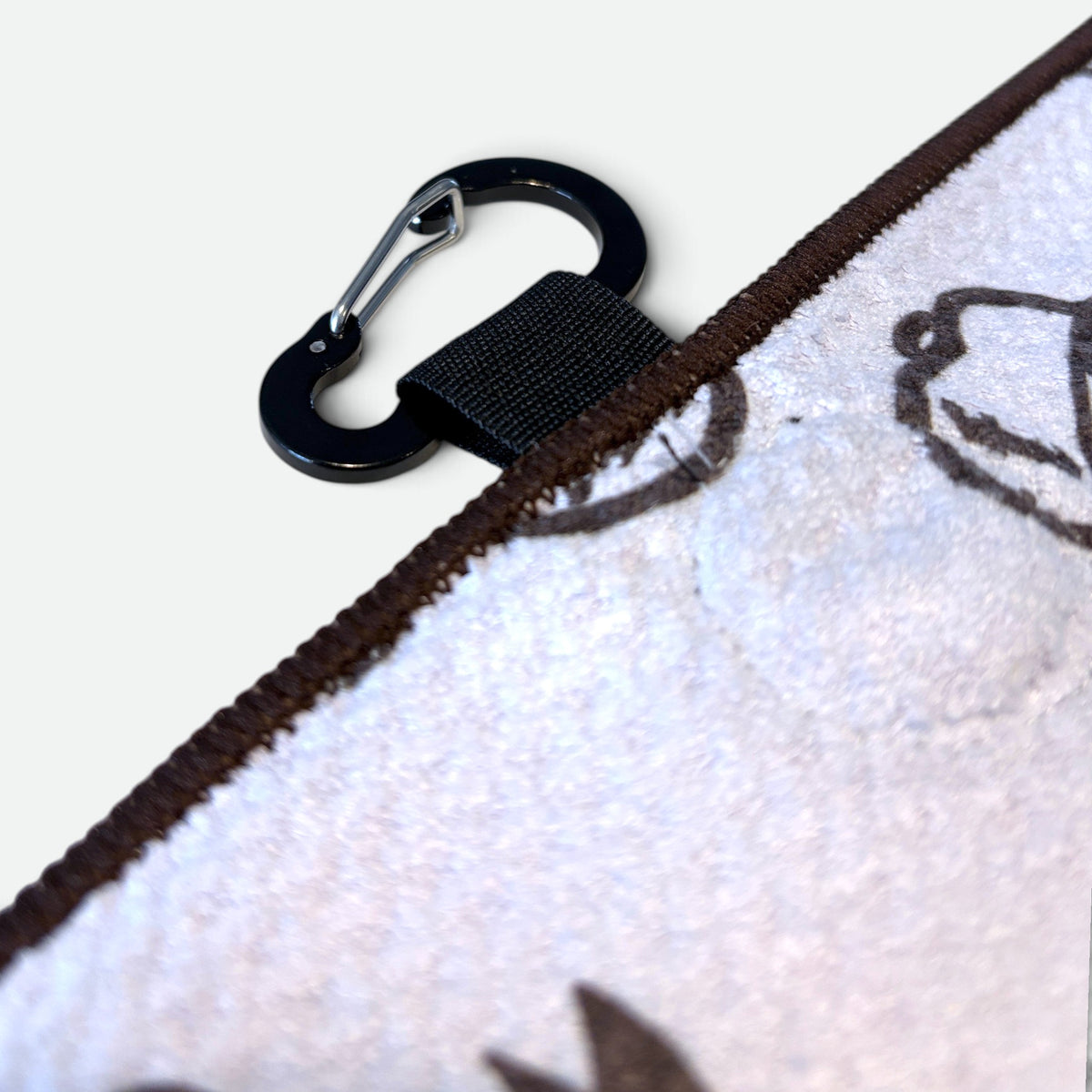 Close-up of a white waffle golf towel with a dark brown embroidered border and a black carabiner clip attached by a black strap, on a light grey background.