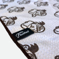 A close-up of a waffle golf towel with a white textured surface, featuring a repeating brown flushed golf mascot pattern and a stitched edge. A black fabric label sewn onto the blanket reads 'Flushed' in white script.