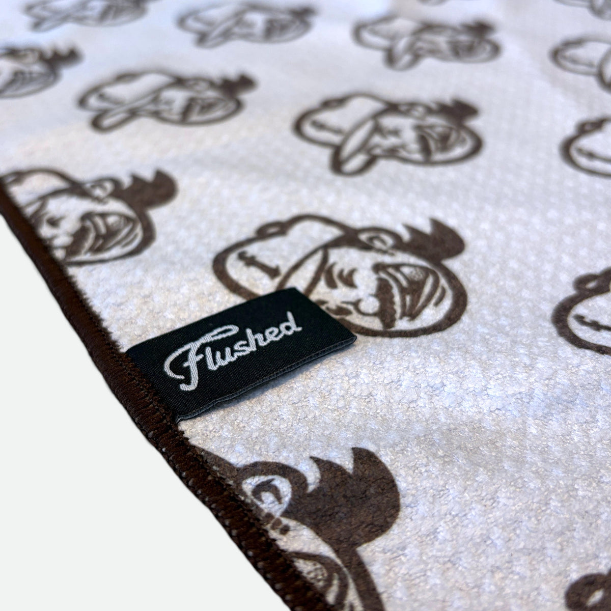 A close-up of a waffle golf towel with a white textured surface, featuring a repeating brown flushed golf mascot pattern and a stitched edge. A black fabric label sewn onto the blanket reads 'Flushed' in white script.