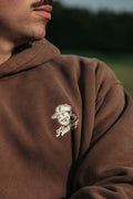A close-up of a person wearing a brown hoodie with a white embroidered graphic on the left chest. The graphic shows a cartoon face with a mullet and moustache wearing a cap with the words 'Flushed golf'.