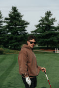 A man wearing a brown hoodie, black sunglasses, and a white golf glove stands on a golf course holding a club.