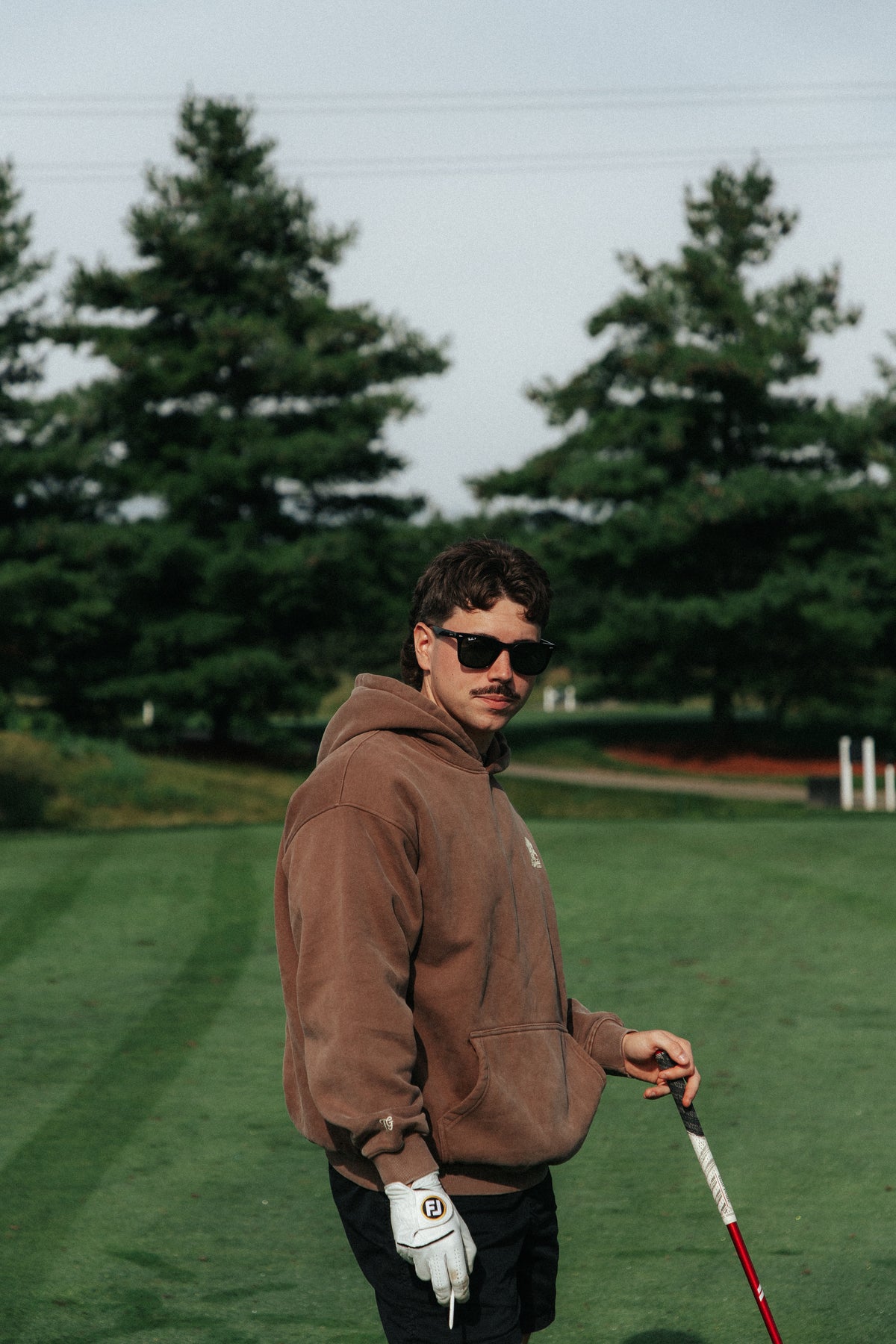 A man wearing a brown hoodie, black sunglasses, and a white golf glove stands on a golf course holding a club.