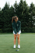 A woman wearing a green hoodie, white skirt, and white sneakers is putting on a golf green with a tree line in the background.