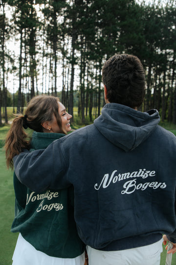 Two people standing outdoors on a golf course, seen from behind, wearing matching dark green and navy hoodies with the words 'Normalize Bogeys' printed in white script across the back.