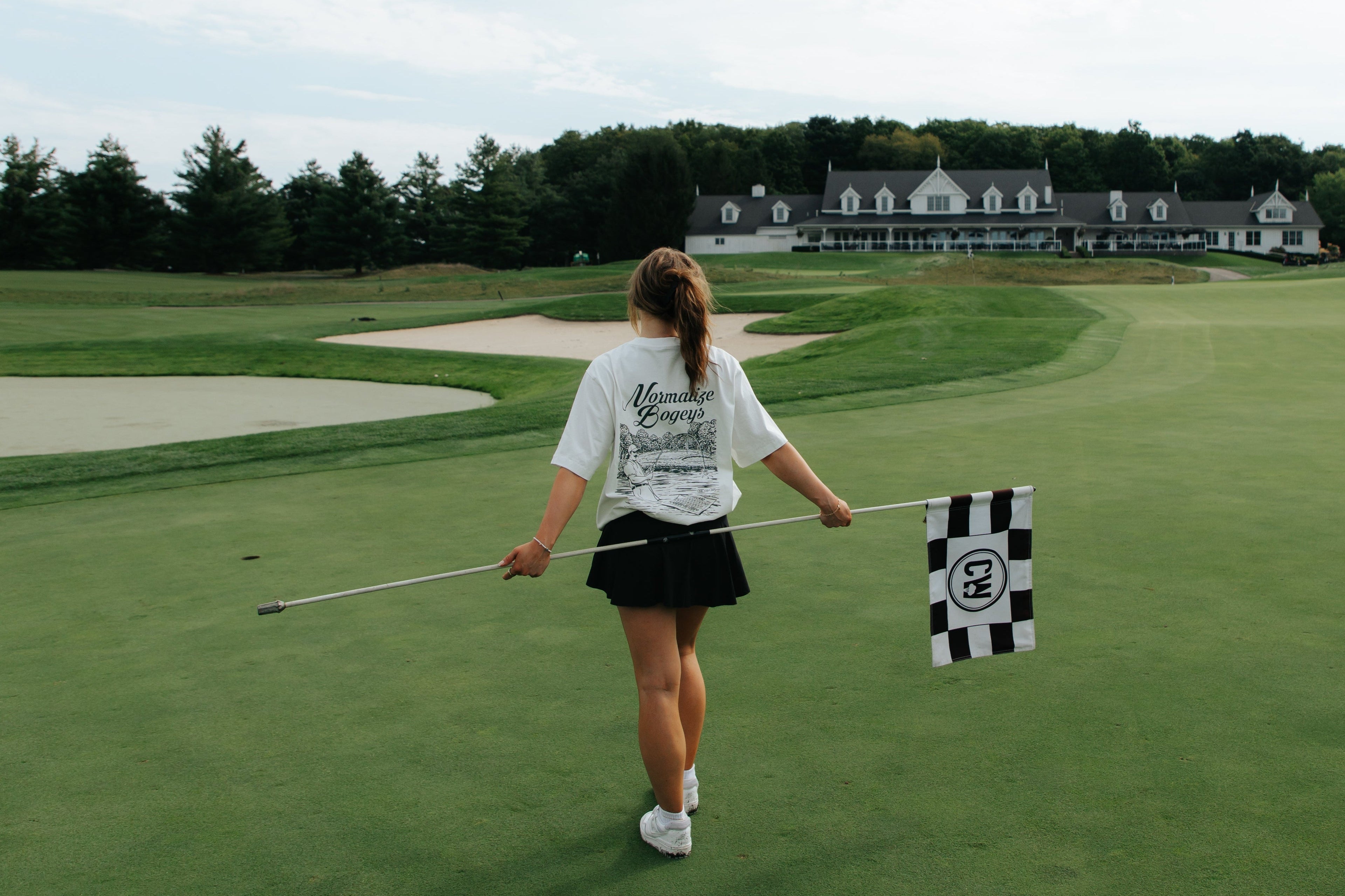 A woman on a golf course holds a checkered flagstick while walking toward a clubhouse in the distance under a cloudy sky.