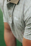Close-up of a person wearing a short-sleeve patterned country golf polo shirt with a beige cactus pattern, featuring a white script logo on the chest.