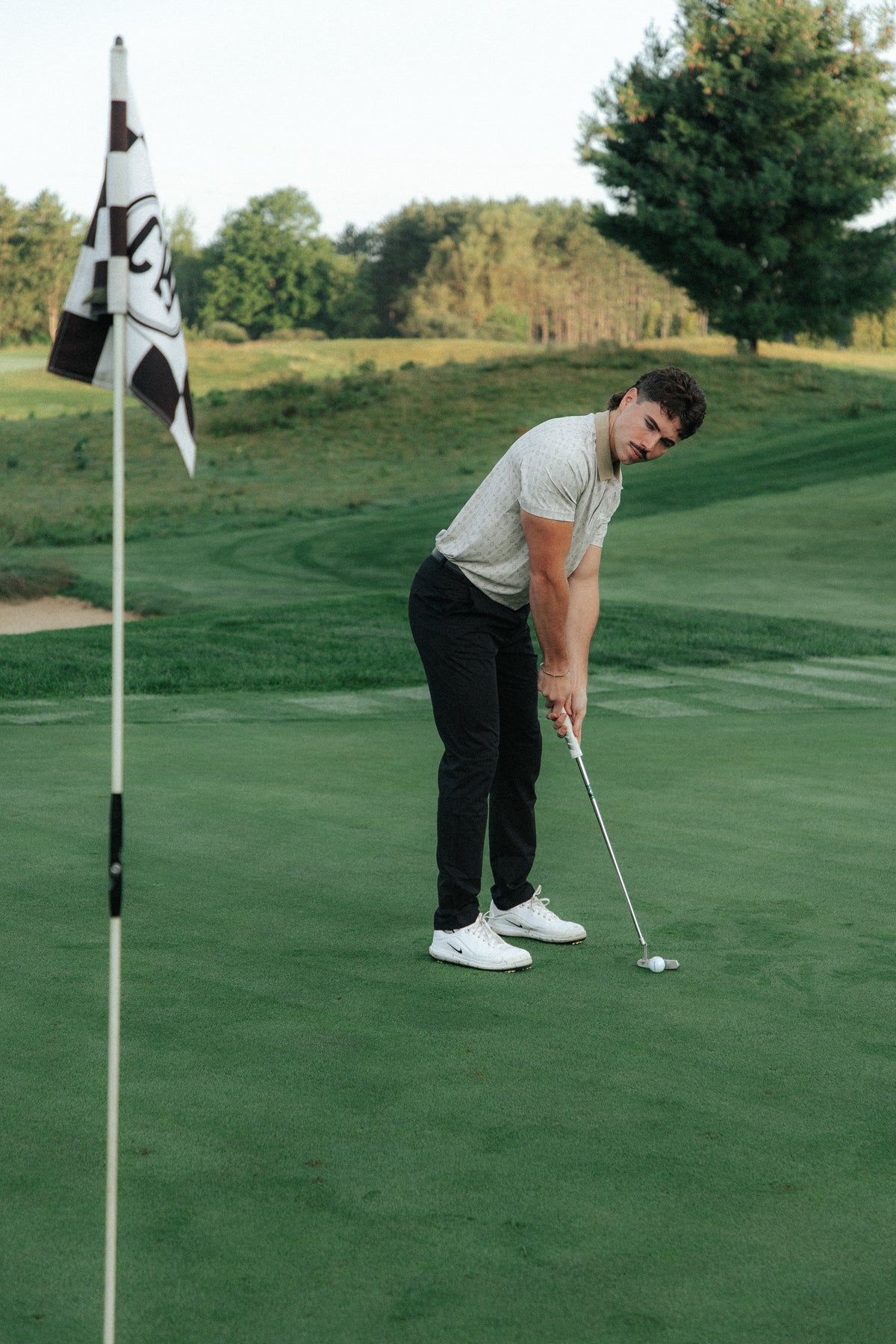 A man on a golf green wearing a tan patterned polo, black pants, and white sneakers, holding a golf club near the flag. A black and white checkered flag is visible in the foreground on a lush green course.