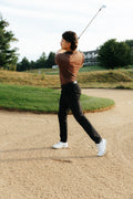 A man in a brown country golf polo shirt, black pants, and white golf shoes is captured mid-swing on a golf course sand bunker.