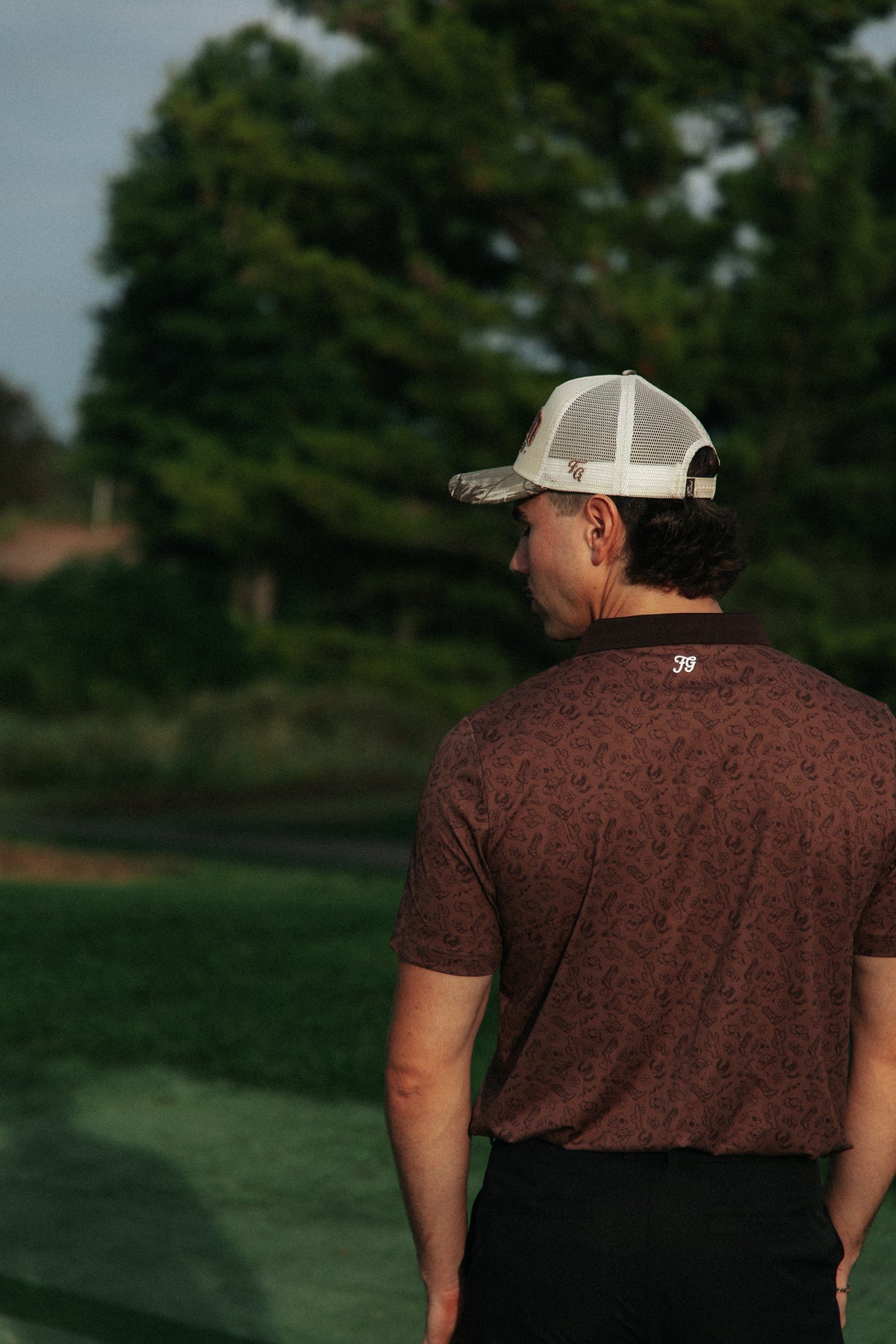 A man on a golf course seen from behind, wearing a brown country patterned short-sleeve golf polo shirt, black pants, and a white trucker cap with a camouflage brim.